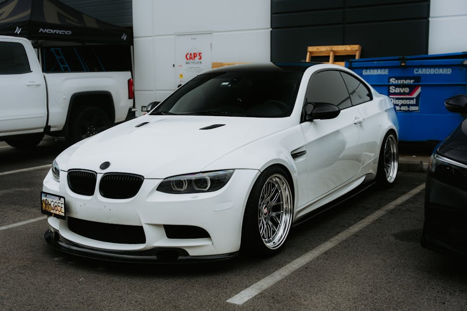 White sports car with tinted windows parked outdoors next to a blue dumpster.