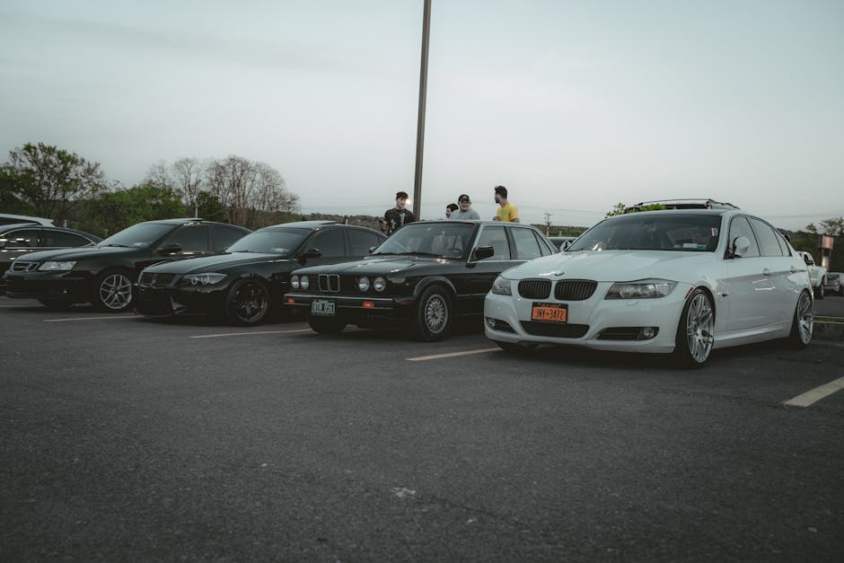 A lineup of BMW cars parked in an outdoor lot with people gathering during the evening.